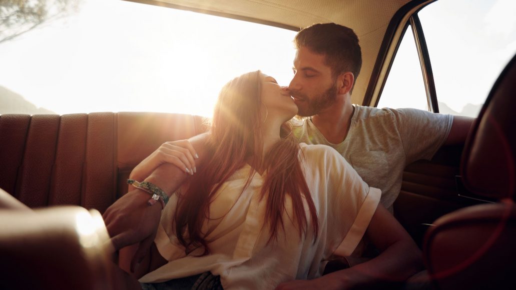 Romantic young couple sitting in back seat of a car and kissing . Affectionate couple on rear seat of a vehicle with sun flare.